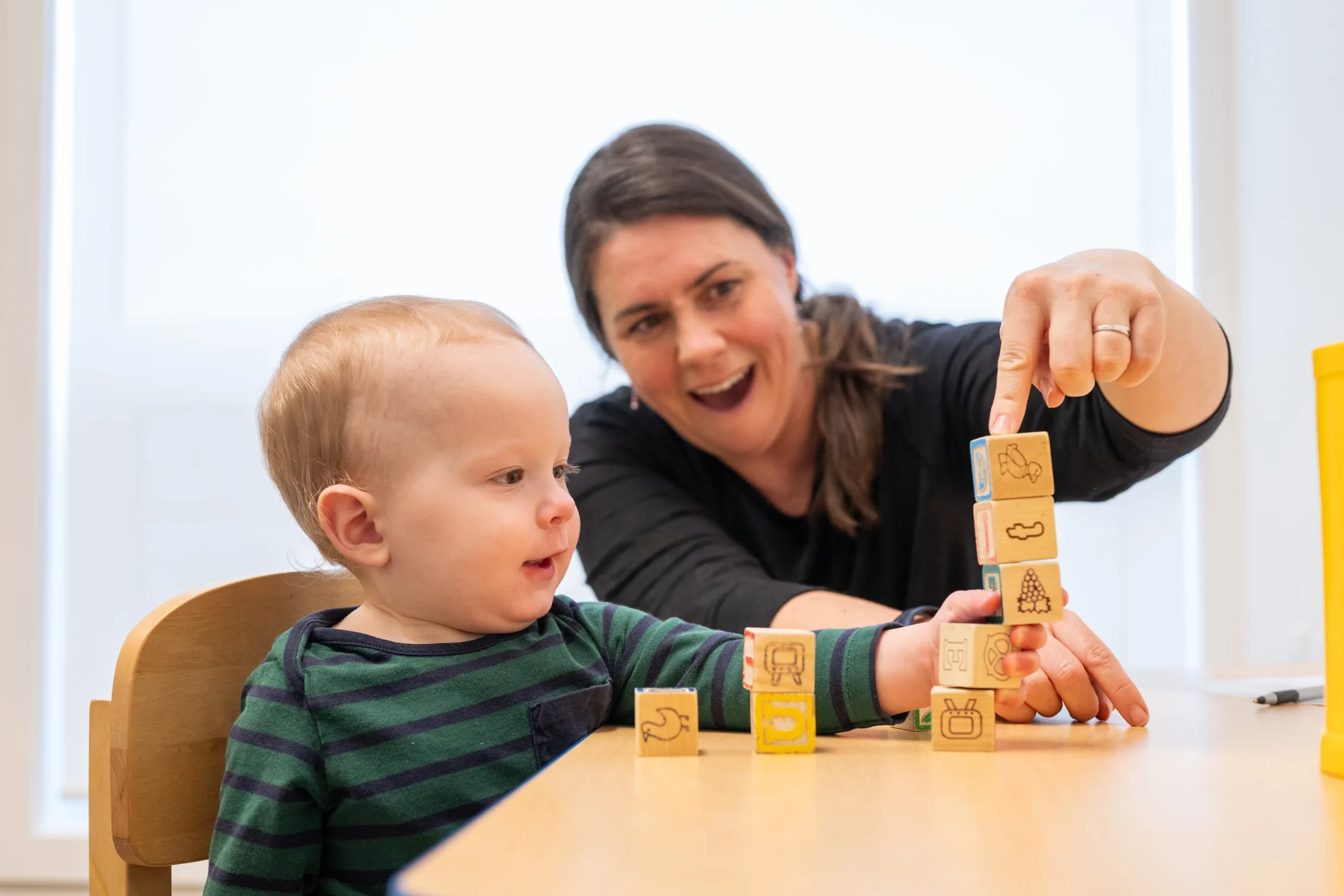 A child is building a tower with blocks while an adult watches and smiles at them from behind.